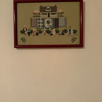 Framed cross-stitch artwork featuring a folk-art style scene of a house with displayed quilts and figures, housed in a reddish-brown...
