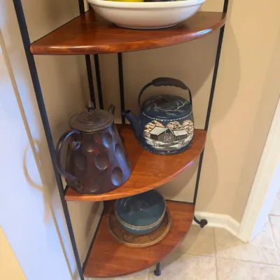 Three-tier wrought iron and wooden corner shelf holding decorative teapots, a bowl with artificial fruit, and ceramic dishware.