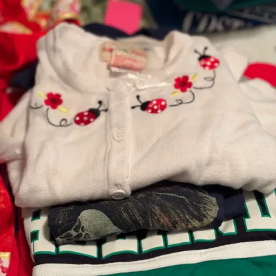 A folded white toddler's cardigan featuring embroidered red ladybugs and flowers along the neckline, resting atop a pile of...