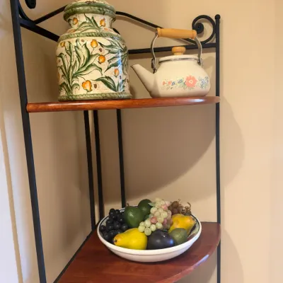Black wrought iron corner baker's rack with wood shelves, holding a hand-painted ceramic jar, small vintage teapot, and a bowl of...
