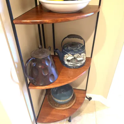 Corner shelving unit with wood tiers and a black metal frame, displaying decorative teapots, a bowl of artificial fruit, and ceramics.