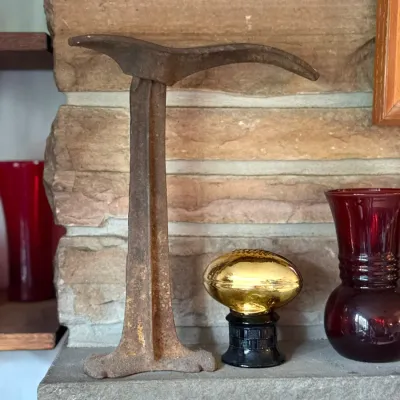 Sale Photo Thumbnail #94: Antique cast iron cobbler's shoe last anvil, displayed on a fireplace mantel alongside a small gold-toned football trophy and ruby red glass vase.