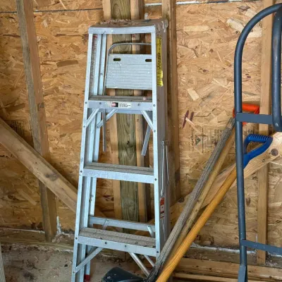 Sale Photo Thumbnail #224: Aluminum stepladder stored in a shed alongside long-handled yard tools and a hand truck.