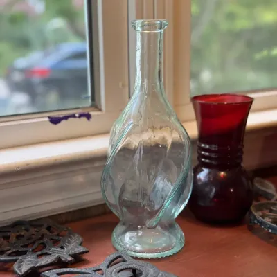 Sale Photo Thumbnail #190: Light green molded swirl glass bottle, displayed on a windowsill alongside cast iron trivets and a ruby red glass vase.