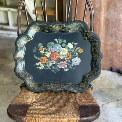 Sale Photo Thumbnail #263: Vintage black toleware serving tray featuring hand-painted floral centerpiece and gilt filigree border, resting on a wooden chair with a woven rush seat.