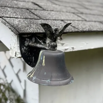 Sale Photo Thumbnail #202: Vintage cast metal dinner bell mounted under an eave, topped with a spread-winged eagle finial and showing weathered patina.