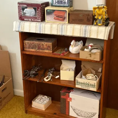 Sale Photo Thumbnail #31: Small three-shelf wooden bookcase displaying various collectibles, including boxed tableware, ceramic swan figurines, a Royal Doulton Bunnykins piece, and a yellow Lego vehicle.
