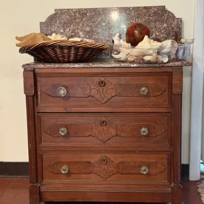 Sale Photo Thumbnail #159: Antique marble-top wooden dresser with three drawers, glass knobs, and carved keyhole accents. Decorative seashells in a woven basket and a glass bowl rest on top.