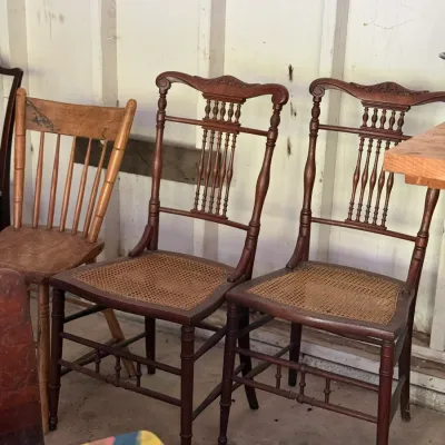 Sale Photo Thumbnail #280: Pair of antique Victorian-style dark wood side chairs with carved crests, turned spindles, and woven cane seats, shown alongside a light wood spindle-back chair.