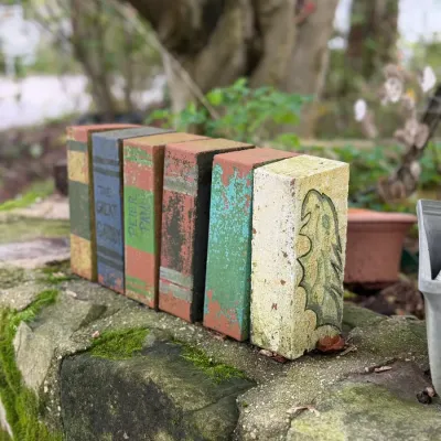 Sale Photo Thumbnail #207: Set of decorative bricks painted as classic book spines, including The Great Gatsby and Peter Pan, showing weathered outdoor wear.
