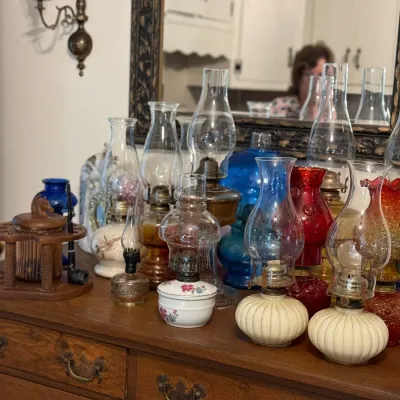 Sale Photo Thumbnail #162: Collection of vintage glass and ceramic oil lamps in various colors and designs, arranged on a wooden dresser alongside a wooden pipe stand.