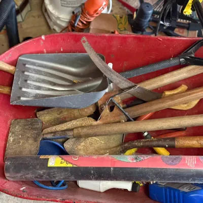 Sale Photo Thumbnail #218: Red metal wheelbarrow filled with assorted hand tools, including shovels, a hand saw, a square edger, and rolled landscape fabric.