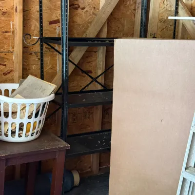Sale Photo Thumbnail #226: Black metal multi-tier shelving unit, accompanied by a small wooden side table, a white laundry basket, and loose building materials in a garage.