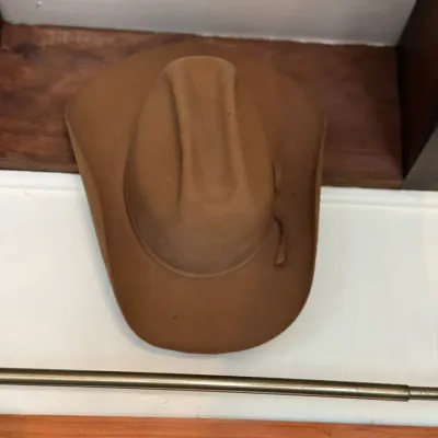 Sale Photo Thumbnail #131: Brown felt cowboy hat resting on a wooden shelf, featuring a traditional cattleman crease and matching hatband.