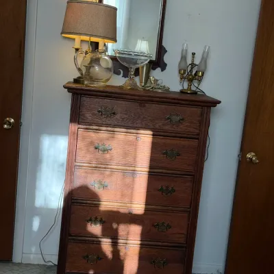 Sale Photo Thumbnail #19: Vintage dark oak five-drawer tall dresser with brass bail handles, topped with decor including a table lamp, glass bowl, and wall mirror.