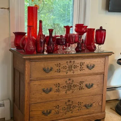Sale Photo Thumbnail #143: Vintage carved oak three-drawer chest featuring ornate floral drawer fronts and bail handles, topped with a large collection of red glass vases, bottles, and tableware.