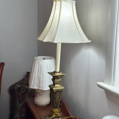 Sale Photo Thumbnail #11: Traditional brass buffet table lamp with a tall candlestick column and cream bell lampshade, displayed on a wooden shelf alongside other vintage lamps.
