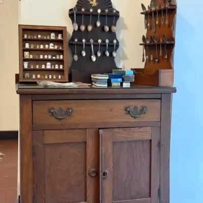 Sale Photo Thumbnail #144: Vintage dark wood dry sink cabinet featuring a top drawer and lower double doors, topped with wall-mounted spoon racks and a shadow box display of souvenir thimbles.