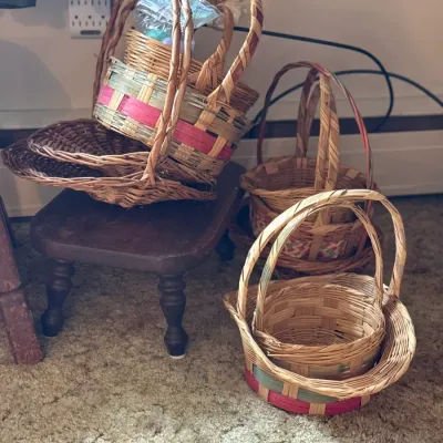 Sale Photo Thumbnail #96: Collection of assorted woven wicker and reed Easter baskets sitting on and around a small dark wood footstool.