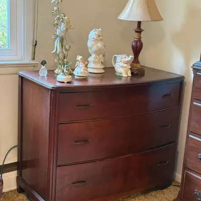 Sale Photo Thumbnail #46: Vintage dark wood bow-front dresser featuring three drawers, topped with various decorative figurines, ceramics, and a table lamp.