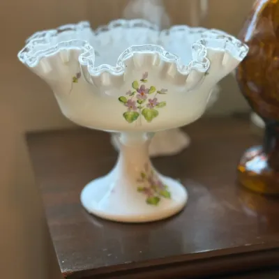 Sale Photo Thumbnail #86: Fenton hand-painted milk glass pedestal compote featuring a ruffled crest rim with silver trim and delicate violet floral motifs.
