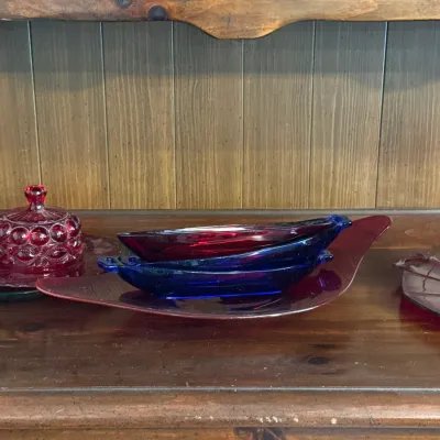 Sale Photo Thumbnail #109: Mid-century colorful glassware, including a red glass leaf platter, cobalt blue and ruby red boat-shaped dishes, and a ruby red Moon and Stars pattern butter dish, resting on a wooden hutch.