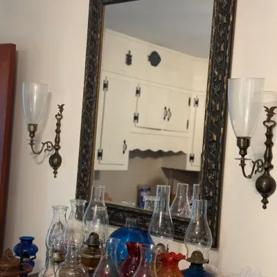 Sale Photo Thumbnail #163: Large dark carved wood mirror flanked by a pair of wall sconces, displayed above a collection of vintage glass oil lamps.
