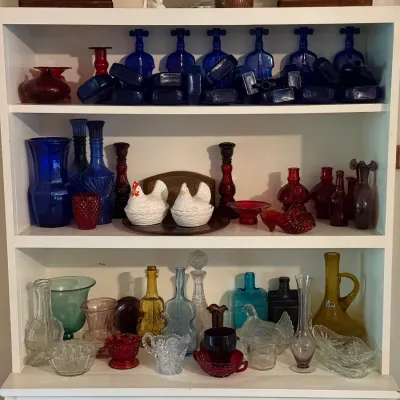 Sale Photo Thumbnail #151: A white painted shelving unit filled with vintage glassware, including cobalt blue bottles, ruby red glass, ceramic hen-on-nest dishes, and various decanters.