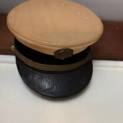 Sale Photo Thumbnail #128: Vintage military service visor cap with a tan fabric crown, dark band, brass insignia emblem, and leather brim.