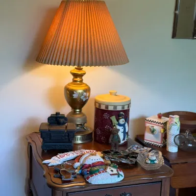 Sale Photo Thumbnail #82: Vintage brass hand-painted table lamp with a pleated shade set atop a wooden nightstand with various winter-themed decor items and knickknacks.