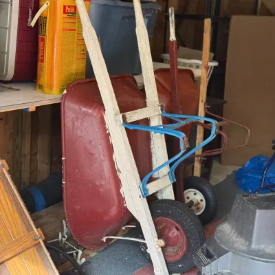 Sale Photo Thumbnail #230: Vintage red metal wheelbarrow with wooden handles, showing signs of heavy use and weathered paint.