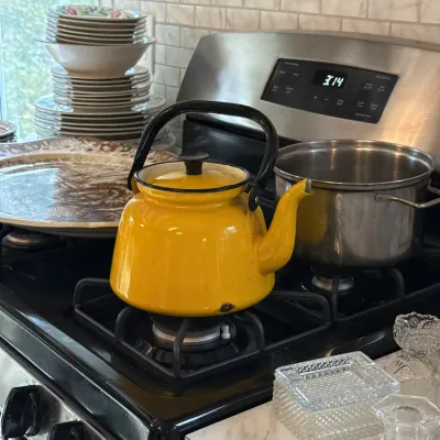 Sale Photo Thumbnail #120: Vintage bright yellow enamel tea kettle with a black handle, resting on a stove next to cookware and stacks of dinnerware.