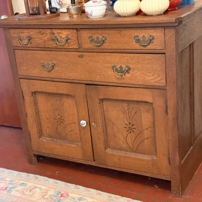 Sale Photo Thumbnail #160: Antique oak server cabinet featuring carved floral panel doors, three upper drawers with brass hardware, and a white ceramic knob.