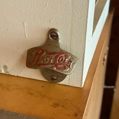 Sale Photo Thumbnail #222: Vintage wall-mounted Pepsi-Cola bottle opener featuring embossed red lettering on a metal surface.