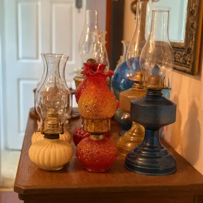 Sale Photo Thumbnail #161: Collection of vintage glass oil lamps displayed on a wooden cabinet, including colorful blue, amber, red patterned, and off-white bases with clear glass chimneys.