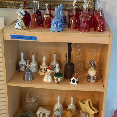 Sale Photo Thumbnail #62: Collection of vintage decorative bells and small figurines arranged on a light wood bookshelf, featuring red glass, blue transferware, and porcelain designs.