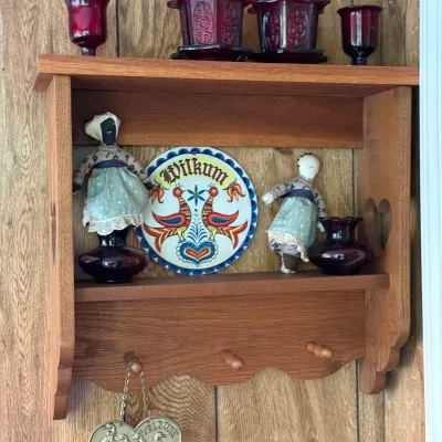 Sale Photo Thumbnail #172: Wall-mounted wooden display shelf with pegs, featuring heart side-cutouts and holding red pressed glass glassware, cloth dolls, and a Pennsylvania Dutch ceramic 'Wilkum' plate.