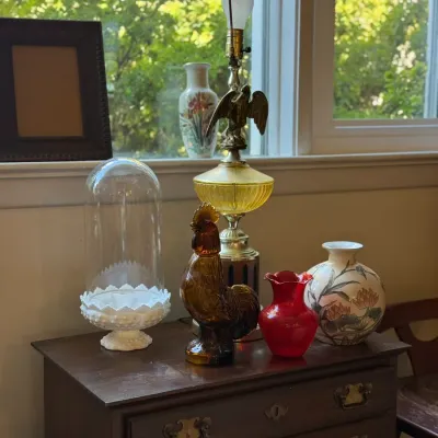 Sale Photo Thumbnail #87: Collection of decorative home accents displayed on a vintage dark wood chest of drawers, including an amber glass rooster figurine, a brass eagle table lamp, milk glass cake stand with glass cloche, and various ceramic and glass vases.