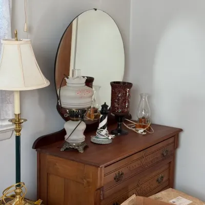 Sale Photo Thumbnail #17: Vintage carved wooden vanity dresser with an attached oval mirror, topped with various decor items including a hand-painted glass lamp and lighthouse figurine.