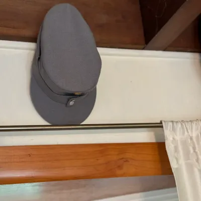 Sale Photo Thumbnail #130: Gray military-style field cap or kepi, featuring a cloth brim and front silver-tone button detail, displayed hanging on a wall.