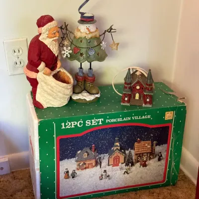 Sale Photo Thumbnail #65: Collection of Christmas decor featuring a Santa Claus figure, a snowman figurine, a miniature illuminated church, and a box for a 12-piece porcelain village set.