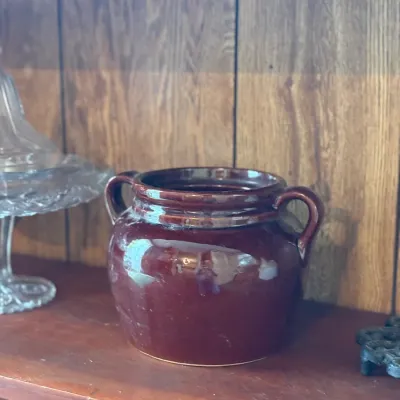 Sale Photo Thumbnail #179: Vintage dark brown glazed stoneware bean pot with dual side handles, resting on a wooden shelf.
