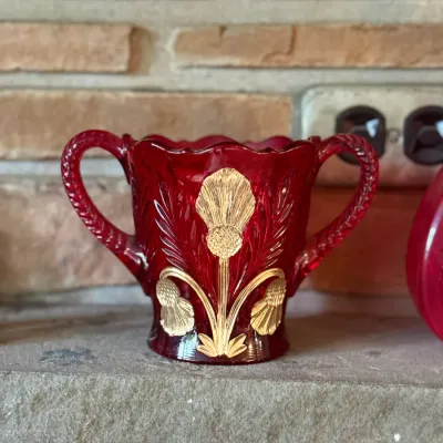 Sale Photo Thumbnail #93: Antique ruby stained glass sugar bowl featuring an embossed gold-gilt thistle pattern with double handles and a scalloped rim.