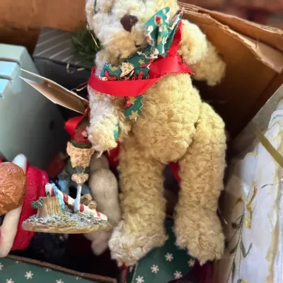 Sale Photo Thumbnail #56: Plush beige teddy bear ornament wearing a patterned bow and red ribbon, packed in a box alongside various holiday decorations.