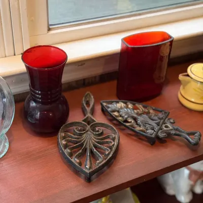 Sale Photo Thumbnail #189: Pair of ornate cast iron trivets resting on a wooden windowsill alongside red glass vases and ceramic cookware.