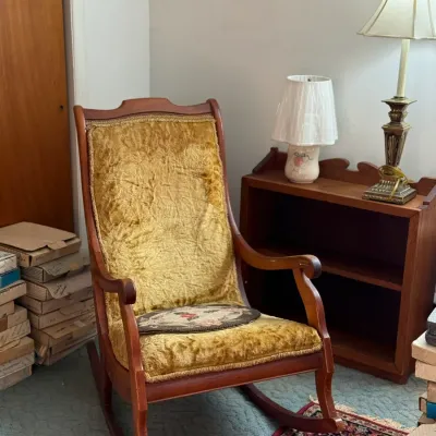 Sale Photo Thumbnail #21: Vintage high-back wooden rocking chair upholstered in mustard yellow crushed velvet, displayed alongside a small wooden bookcase and traditional table lamps.
