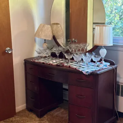 Sale Photo Thumbnail #10: Vintage dark wood vanity desk featuring a large round mirror, seven drawers, and a display of clear crystal stemware set on a runner.