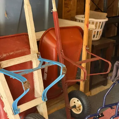 Sale Photo Thumbnail #229: Two vintage wooden-handled hand trucks with metal frames alongside red True Temper wheelbarrows in storage.