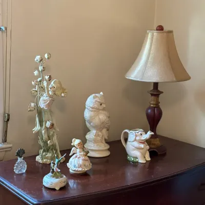Sale Photo Thumbnail #42: Collection of decorative porcelain and ceramic figurines, music boxes, and a small table lamp displayed atop a dark wood dresser.