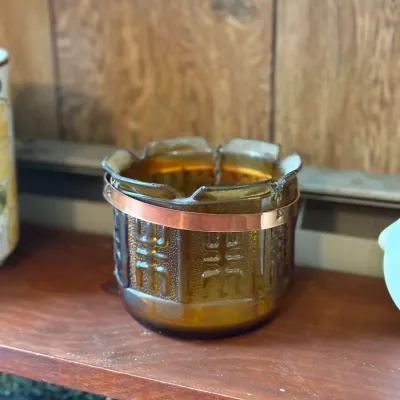 Sale Photo Thumbnail #176: Vintage amber pressed glass votive holder with a scalloped rim, embossed Asian-style characters, and a copper band collar.